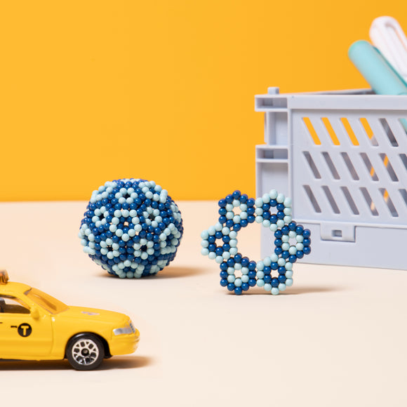 The Speks Elements: Zephyr consists of colorful magnetic balls and shapes in blue and light blue hues. It features a spherical design and a flower-like structure, perfect for creative play and stress relief. A miniature yellow taxi toy is placed nearby for scale, set against a vibrant yellow background.