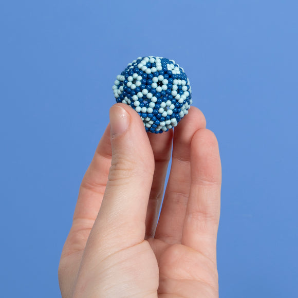 A hand holding a colorful, spherical fidget toy made of hundreds of small beads in various shades of blue and white, set against a light blue background.