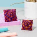 Two colorful fidget toys made of magnetic beads, shaped like small pillows. One features a vibrant pattern of purple and orange beads, while the other has a similar design with a spiral effect. They are placed on a light gray surface next to pink lined paper and colorful pens.