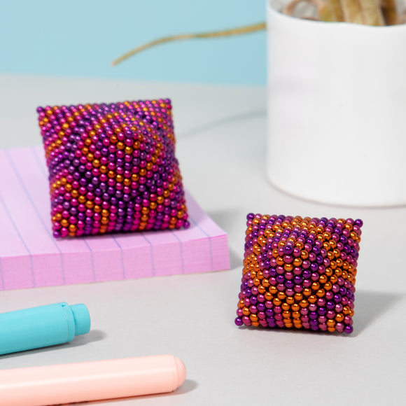 Two colorful fidget toys made of magnetic beads, shaped like small pillows. One features a vibrant pattern of purple and orange beads, while the other has a similar design with a spiral effect. They are placed on a light gray surface next to pink lined paper and colorful pens.