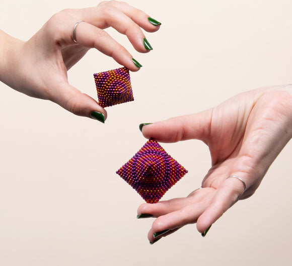 Two hands demonstrating the Speks Energize Gradient, featuring vibrant purple and red magnetic beads shaped into cube-like structures. One hand holds a smaller version while the other showcases a larger one, highlighting their playful, tactile design.