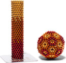 A colorful set of magnetic beads from the Speks Ignite Gradient collection, featuring a tall stack of beads transitioning from golden yellow to deep red, alongside a sphere of similar beads. Both are displayed on a clear base, emphasizing their vivid gradient colors.