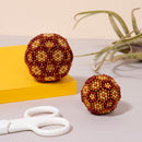 A colorful display of two textured beaded spheres in shades of red and gold, sitting beside a yellow notepad and white scissors. The larger sphere features intricate bead patterns, while the smaller one has a simpler design. An air plant is subtly positioned in the background, adding a touch of greenery to the scene.