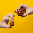 Two hands holding Speks Ignite Gradient fidget balls against a vibrant yellow background. The larger ball features a gradient of metallic red, orange, and gold speks, while the smaller ball exhibits a similar color scheme in a diminutive size, showcasing their fun and engaging design.