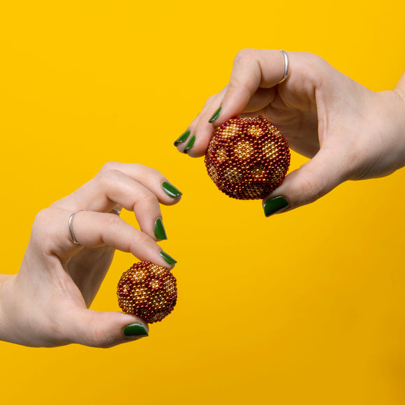 Two hands holding Speks Ignite Gradient fidget balls against a vibrant yellow background. The larger ball features a gradient of metallic red, orange, and gold speks, while the smaller ball exhibits a similar color scheme in a diminutive size, showcasing their fun and engaging design.