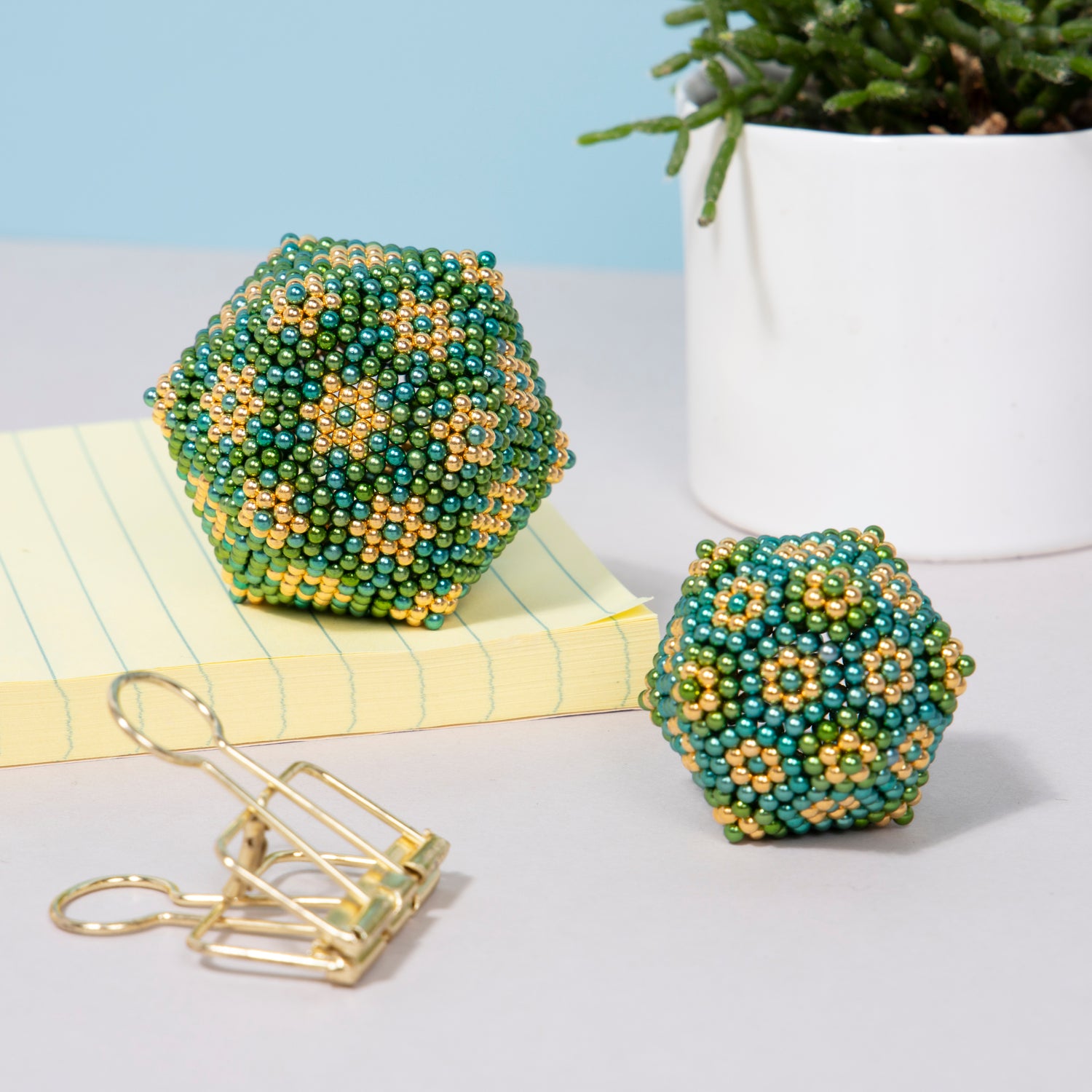 The Speks Inspire Gradient is a vibrant, beaded fidget toy featuring intricate floral designs in green and gold hues. The image shows two hexagonal shapes alongside a notepad and a gold binder clip, highlighting its decorative and playful appeal.