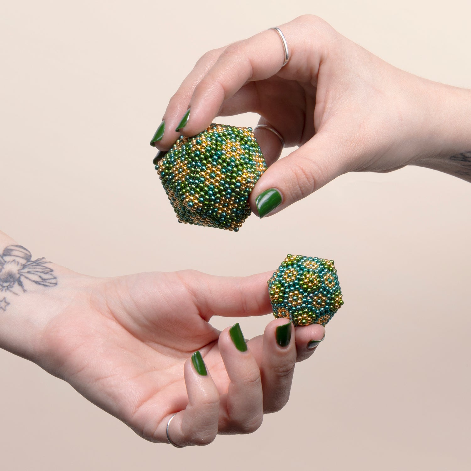 Two hands hold colorful Speks Inspire Gradient, a tactile fidget toy made of magnetic beads. The larger piece is hexagonal with green, gold, and blue patterns, while the smaller one mimics a similar style. Perfect for stress relief and brain stimulation.