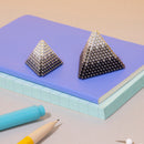 The Speks Reflect Gradient consists of two pyramid-shaped magnetic sculptures made from a gradient of shiny silver and black spheres, displayed on a light purple notebook. Nearby are a light blue marker and a yellow and white pencil, creating a vibrant and creative workspace.