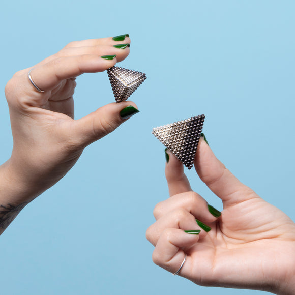 Two hands holding different sizes of Speks Reflect Gradient, a magnetic fidget toy. The toy features small metallic spheres forming pyramid shapes, showcasing a gradient effect.