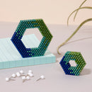Two hexagonal shapes made of colorful magnetic balls sitting on a light blue notepad. A larger hexagon in gradient shades of green and blue is on the left, with a smaller one beside it. White push pins are scattered in the foreground, and a green plant stem is visible in the background.