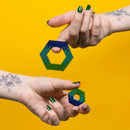 Two hands holding colorful hexagonal shapes made of magnetic beads, showcasing a gradient of blue and green hues. The background is bright yellow, creating a vibrant contrast.