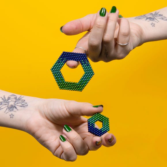 Two hands holding colorful hexagonal shapes made of magnetic beads, showcasing a gradient of blue and green hues. The background is bright yellow, creating a vibrant contrast.