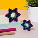 The Speks Unwind Gradient features two colorful star-shaped fidget toys made of magnetic beads, displayed alongside a notepad and colored markers. The larger star is adorned with shades of purple, blue, and green, highlighting its intricate design, while a smaller star lies in front of it. A potted plant adds a touch of greenery to the vibrant yellow background.