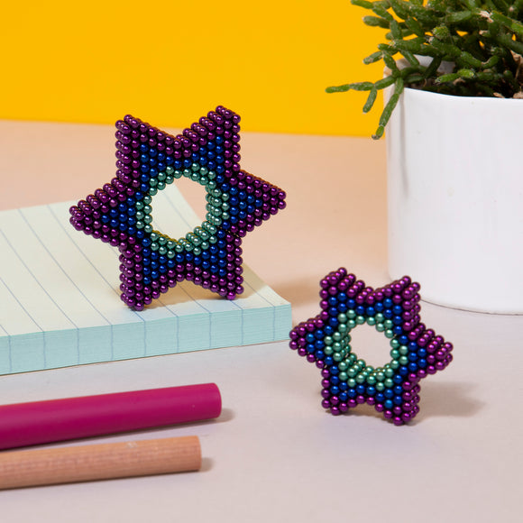 The Speks Unwind Gradient features two colorful star-shaped fidget toys made of magnetic beads, displayed alongside a notepad and colored markers. The larger star is adorned with shades of purple, blue, and green, highlighting its intricate design, while a smaller star lies in front of it. A potted plant adds a touch of greenery to the vibrant yellow background.