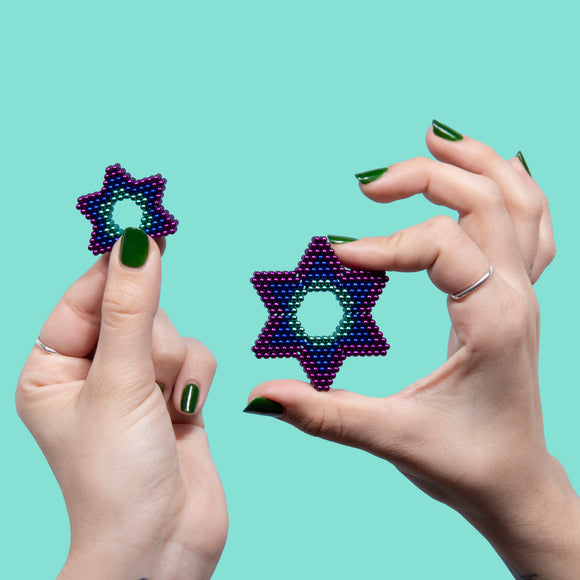 Two hands holding colorful star-shaped fidget toys made of small magnetic spheres. One star is larger and vibrant with gradient colors, while the other is smaller and shares a similar design. The background is a soothing mint green, emphasizing the playful nature of the toys.