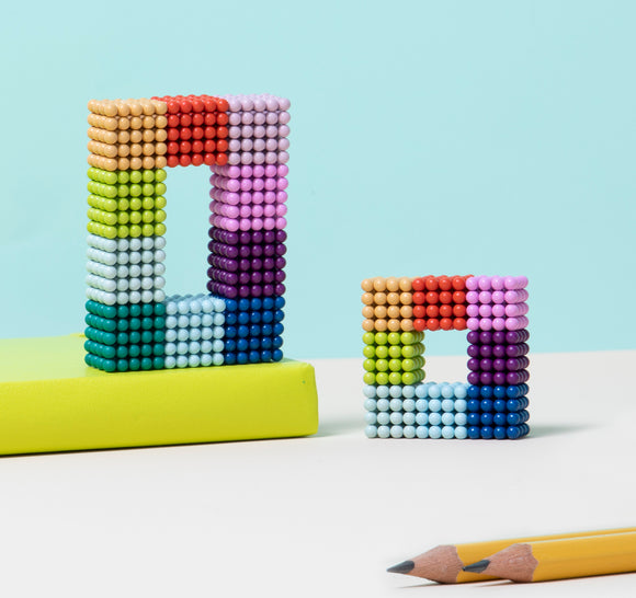 Two colorful structures made of small magnetic beads, with a vibrant gradient effect. The larger one forms a rectangle, and the smaller is a square. A yellow book is in the background, alongside a sharpened pencil.