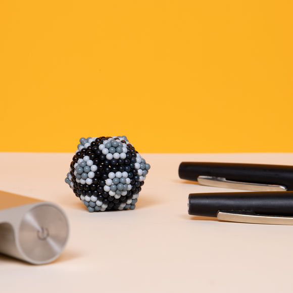 The Speks Pixel Game Over is a colorful fidget toy made from magnetic specks that can be shaped into various designs. The toy features a cluster of interconnected black, gray, and blue magnets, surrounded by stationary pens and a sleek silver item, all against a vibrant yellow background.