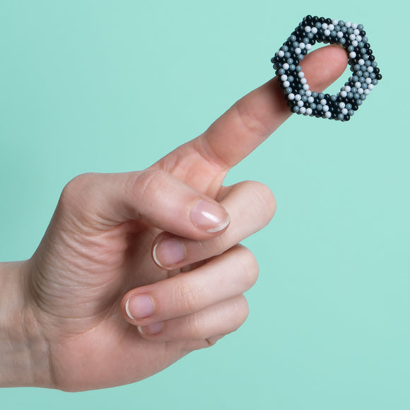 A hand holding a Speks Pixel Game Over fidget toy, which is shaped like a hexagonal ring made of colorful magnetic balls, set against a mint green background.