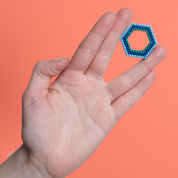 A hand holding a colorful hexagonal fidget toy made of magnetic beads. The toy is set against a vibrant orange background, showcasing its structure and design.