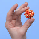 A hand holds the Speks Pixel 1 Up, a colorful fidget toy made of magnetic plastic spheres. The toy forms a hexagonal shape with orange, red, and cream colors, providing a tactile experience and engaging design for stress relief.