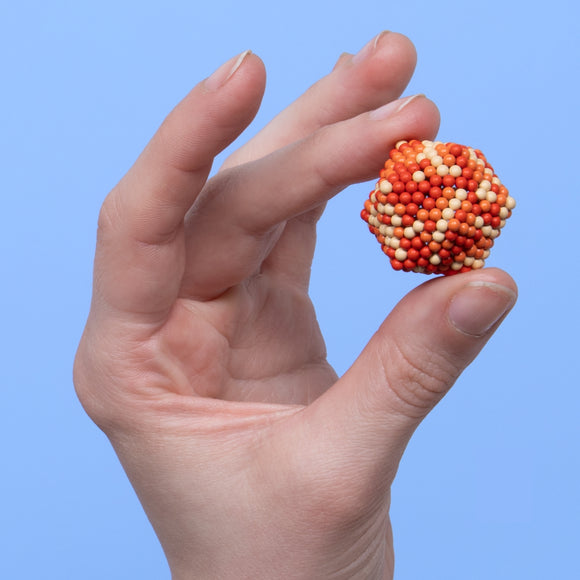 A hand holds the Speks Pixel 1 Up, a colorful fidget toy made of magnetic plastic spheres. The toy forms a hexagonal shape with orange, red, and cream colors, providing a tactile experience and engaging design for stress relief.