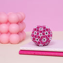 The Speks Pixel Cheat Code is a vibrant, multi-colored sensory toy featuring a spherical design made of small, round magnets in pink and white hues. It rests on a pink notebook with a textured cover, against a soft pink backdrop. The product invites tactile engagement and creative play, offering endless possibilities for imaginative construction and stress relief.