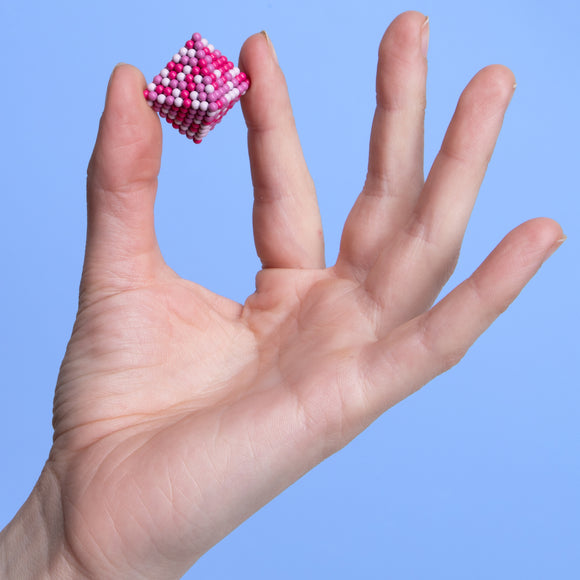 A hand is holding a colorful, cube-shaped fidget toy made of tiny magnetic beads, featuring a pink and white design against a blue background.
