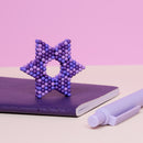 The Speks Pixel 8 Bit is a creative tactile fidget toy made up of small colorful magnetic balls arranged in a star shape, resting on a purple notebook and beside a purple marker. The toy encourages imaginative play and stress relief with its vibrant purple hues.