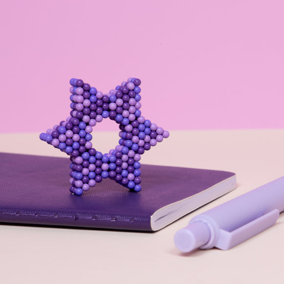The Speks Pixel 8 Bit is a creative tactile fidget toy made up of small colorful magnetic balls arranged in a star shape, resting on a purple notebook and beside a purple marker. The toy encourages imaginative play and stress relief with its vibrant purple hues.