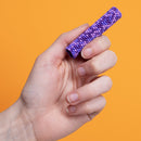 A hand holding a purple Speks Pixel 8 Bit toy, showcasing its rounded, textured surface against a bright orange background. The item is colorful and engaging, designed to provide tactile stimulation and relaxation.
