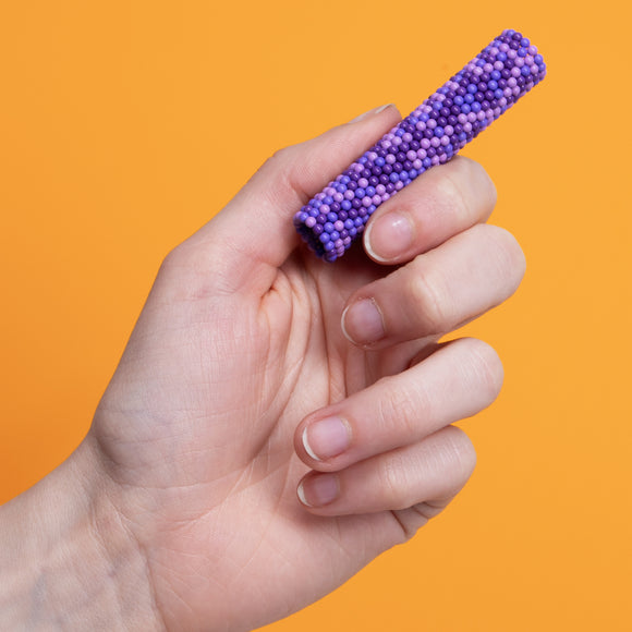 A hand holding a purple Speks Pixel 8 Bit toy, showcasing its rounded, textured surface against a bright orange background. The item is colorful and engaging, designed to provide tactile stimulation and relaxation.
