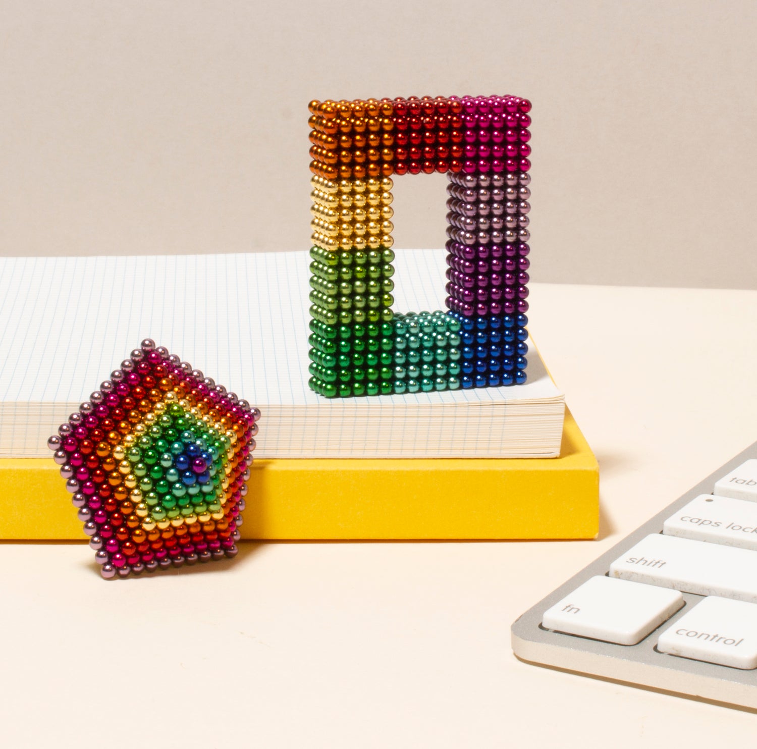 Colorful magnetic spheres arranged into a green and purple rectangular structure with a rainbow-colored hexagonal shape nearby, set against a backdrop of a notepad and a yellow block.