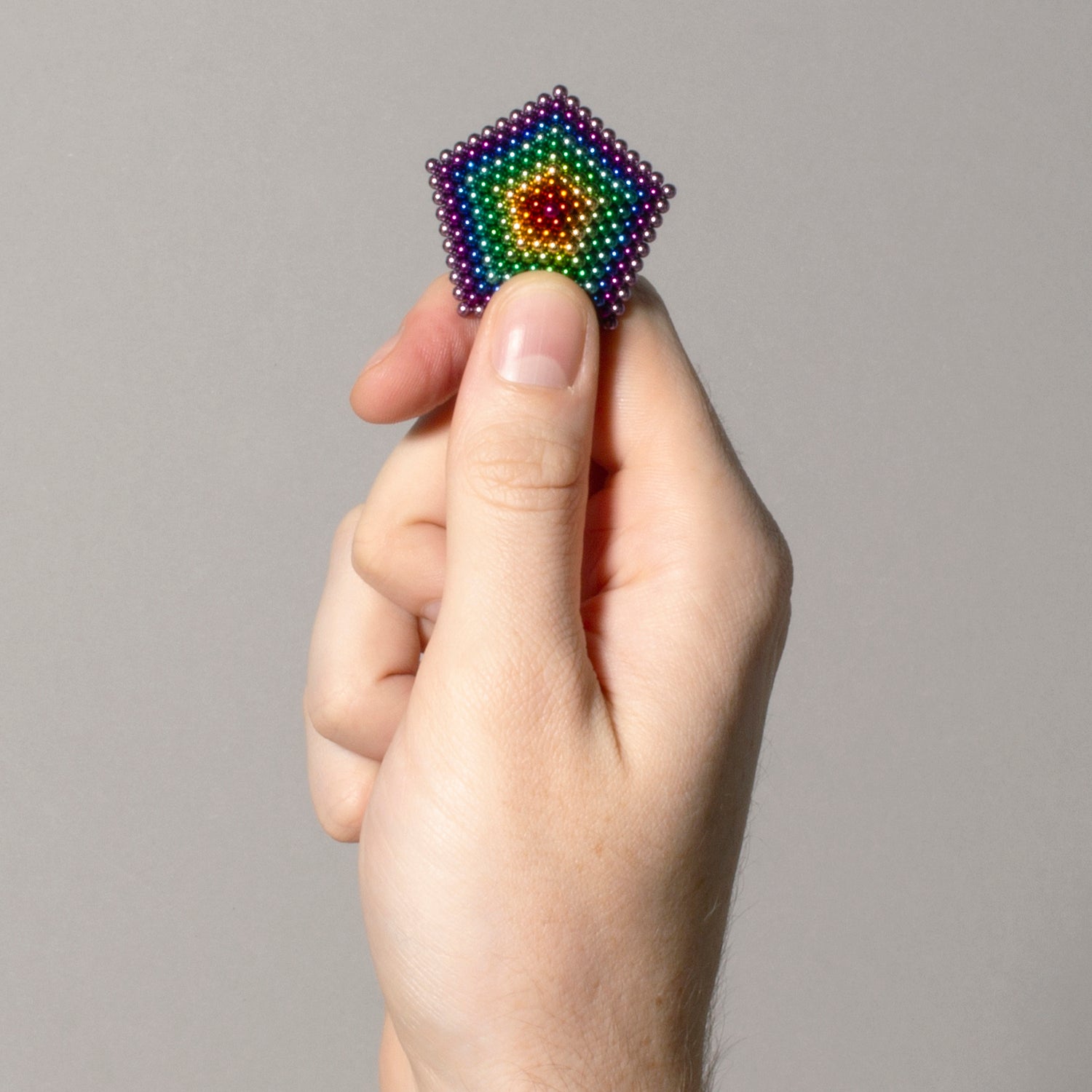 A close-up of a hand holding a colorful hexagonal magnetic ball construction, showcasing the vibrant spectrum of colors and the intricate geometric pattern formed by the small spheres. The background is a neutral gray, highlighting the product details.