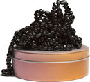 A collection of shiny black beads is artistically arranged atop a pastel-colored tin. The beads vary in shape, creating an intriguing texture and visual appeal. The tin is round with a smooth, metallic finish, contributing to the overall aesthetic of the display.