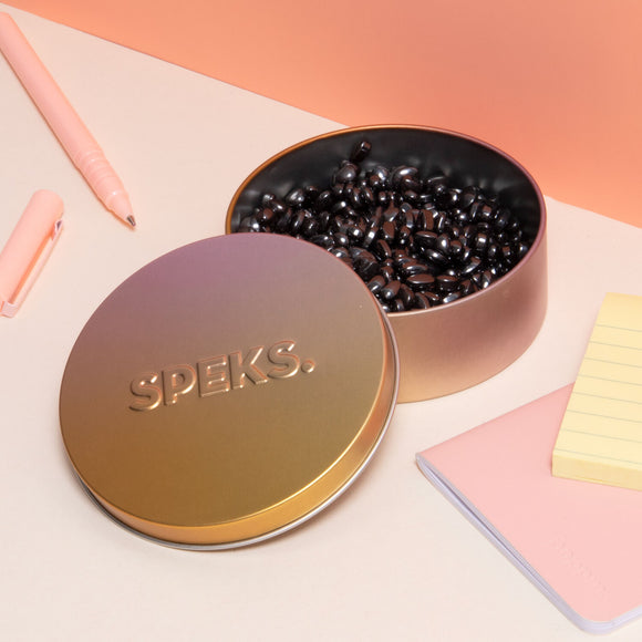 A stylish metal tin with the embossed logo 'SPEKS' on the lid, partially open to reveal glossy black beads inside. The tin is placed on a light-colored surface alongside a pink pen, a pink notepad, and yellow sticky notes. The background features a soft gradient in pastel peach tones, creating a modern and inviting workspace aesthetic.