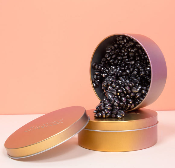 A colorful tin container with a lid slightly ajar, spilling out shiny black bead-like objects. The background features a soft, peachy pink hue, enhancing the product's visual appeal.