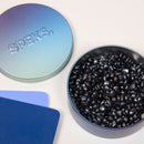 A round, gradient-colored tin labeled 'SPEKS' containing shiny black fidget beads. The tin is partially open, revealing the glossy beads inside. Beside the tin are two colorful cards in blue and lavender, each with smooth edges, set against a beige background.