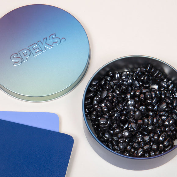 A round, gradient-colored tin labeled 'SPEKS' containing shiny black fidget beads. The tin is partially open, revealing the glossy beads inside. Beside the tin are two colorful cards in blue and lavender, each with smooth edges, set against a beige background.
