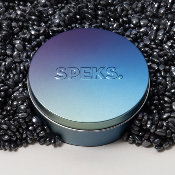 A sleek, gradient metallic tin labeled 'SPEKS.' surrounded by a mass of shiny black beads. The tin features a blue to green ombre finish, adding a modern touch to the serene design. Ideal for storing fidget or sensory items.