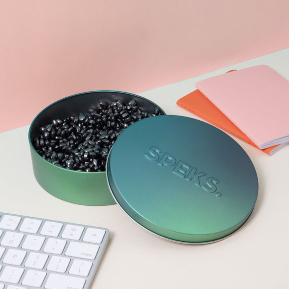 A stylish, round metallic container from SPEKS, showcasing an open lid revealing an assortment of small black spherical magnets inside. The background features a light pastel pink wall, along with a white keyboard and colorful notebooks in pink and orange, creating a modern workspace aesthetic.