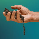 A hand holding a bundle of small, smooth, black stones, with some stones cascading down the fingers. The background is a gradient of soft teal, creating a calming atmosphere that complements the sensory experience of the product.