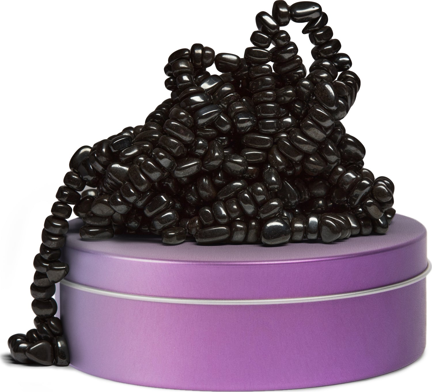 A colorful round tin in purple with a metallic band sits on a white surface, overflowing with a cluster of shiny black beads. The beads vary in size and shape, creating a textured appearance as they drape over the edges of the tin.