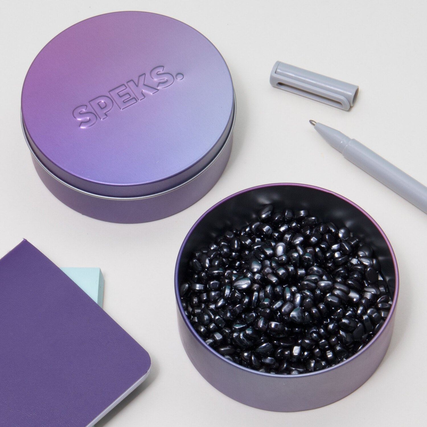 SPEKS Crags: Vitality features a sleek, purple metallic tin containing a collection of shiny black fidget beads. The tin is surrounded by a light gray pen, a notebook, and sticky notes, showcasing a contemporary and organized workspace.