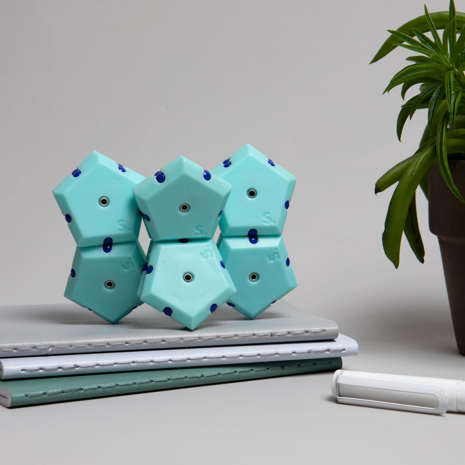 A colorful Speks Geode: Mile High set featuring interconnected hexagonal magnets in a soothing mint color with dark blue accents. The playful design showcases its versatility, sitting on top of a stack of notebooks beside a small potted plant.