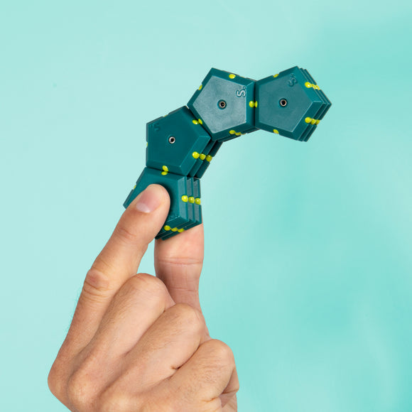 A person holds a uniquely shaped teal and yellow Speks Geode: Monstera puzzle, showcasing its flexible segments. The background is a soft mint green, enhancing the visual appeal of the product.