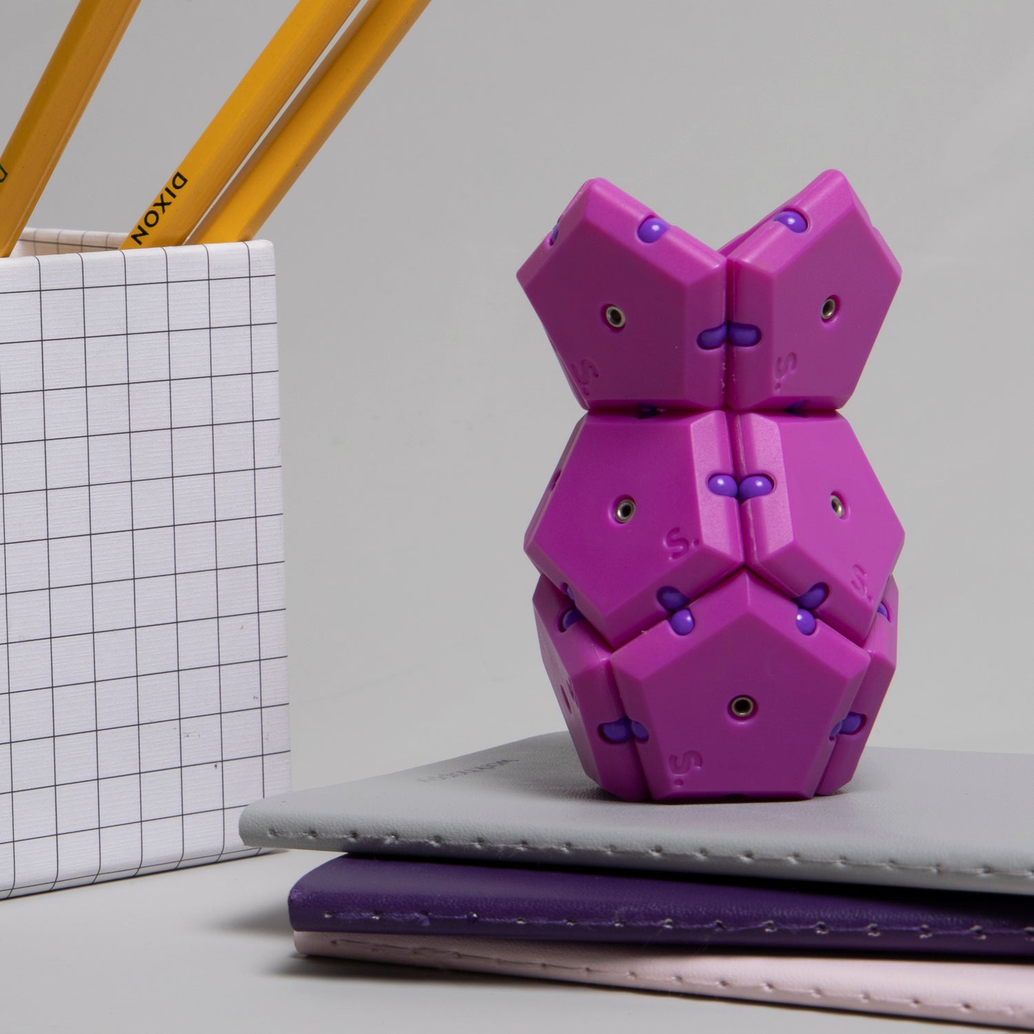 The Geode Razzle Dazzle by Speks is a vibrant pink and purple mechanical puzzle featuring hexagonal shapes that can be stacked and aligned in various configurations. It sits atop a stack of notebooks and next to a pencil holder, showcasing its playful and engaging design. This item is perfect for enhancing focus and creativity, making it an ideal fidget toy or desk accessory.