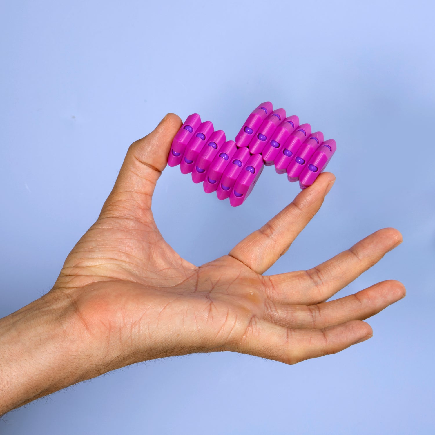 A person's hand holding the Geode Razzle Dazzle, a colorful, flexible fidget toy consisting of interlocking segments that can bend and twist. The toy features a vibrant purple color with blue accents and is set against a light blue background.