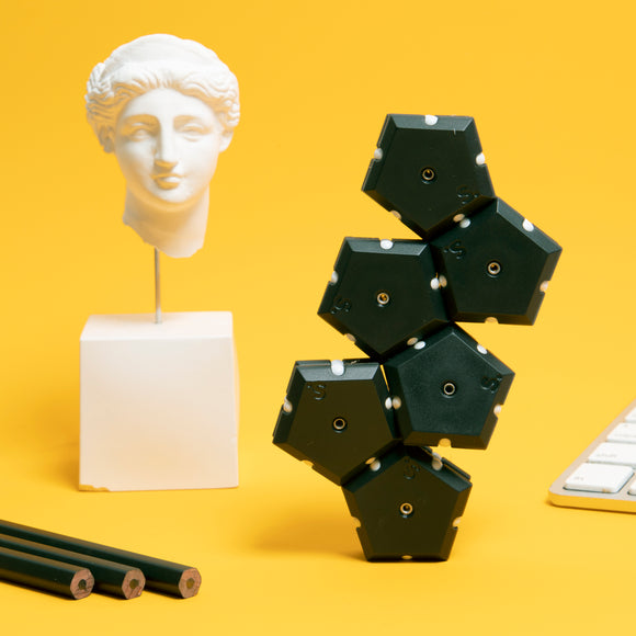 The Speks Geode: Snake Eyes features a playful design with black hexagonal pieces stacked creatively against a bright yellow background. Nearby, there is a white bust statue, and three green pencils are positioned in front of a keyboard, adding to the artistic display.