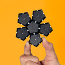 A hand holding the Speks Geode: Snake Eyes, a unique geometric puzzle made of black hexagonal pieces with white details. The pieces connect magnetically, forming an eye-catching shape. The background is a vibrant orange, enhancing the product's visual appeal.