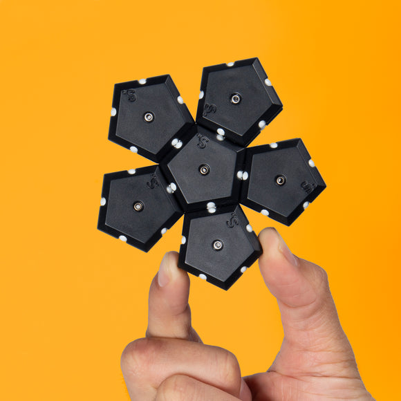 A hand holding the Speks Geode: Snake Eyes, a unique geometric puzzle made of black hexagonal pieces with white details. The pieces connect magnetically, forming an eye-catching shape. The background is a vibrant orange, enhancing the product's visual appeal.